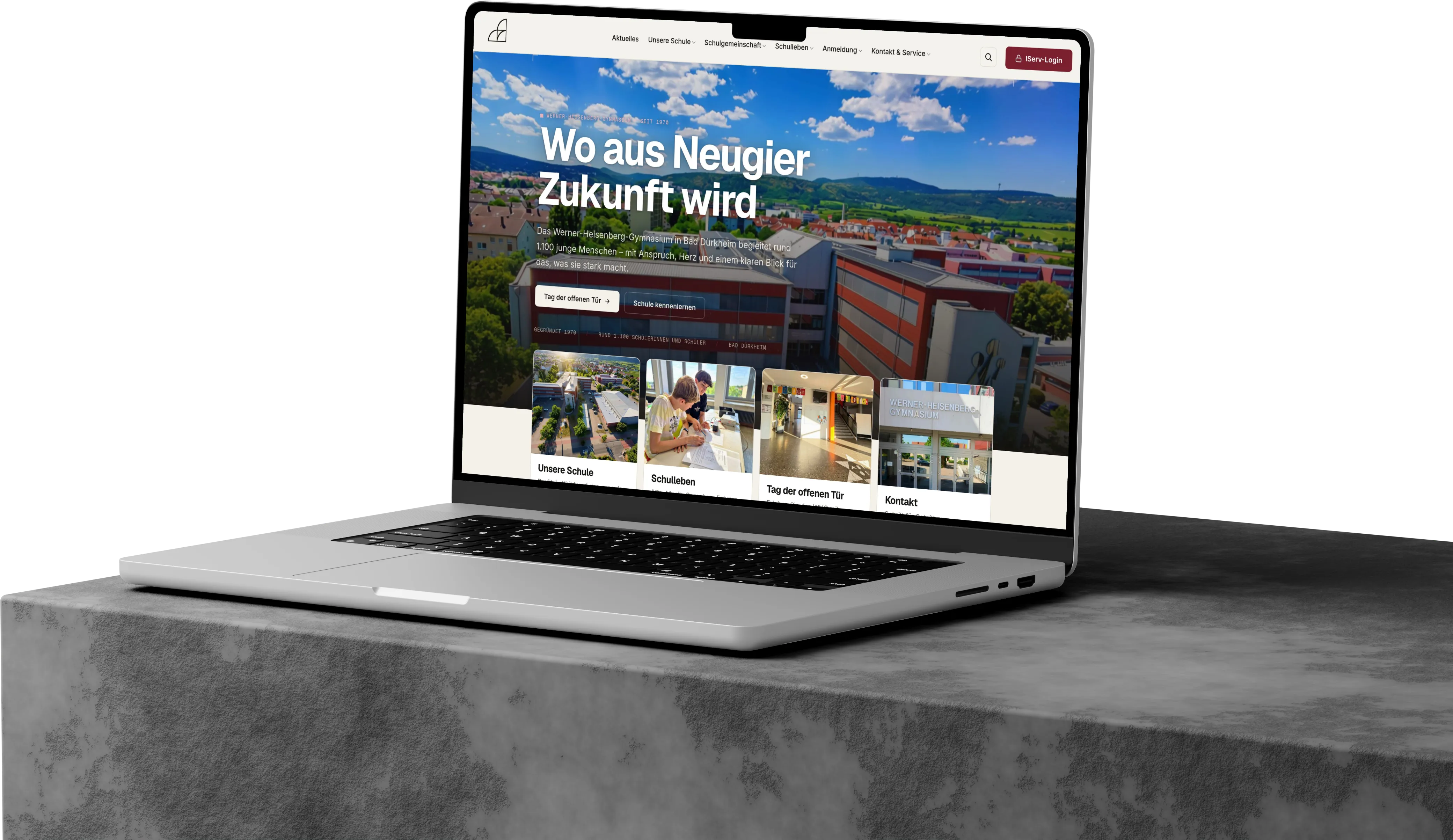 MacBook auf einem Betonsockel mit der Startseite des Werner-Heisenberg-Gymnasiums – „Wo aus Neugier Zukunft wird.“